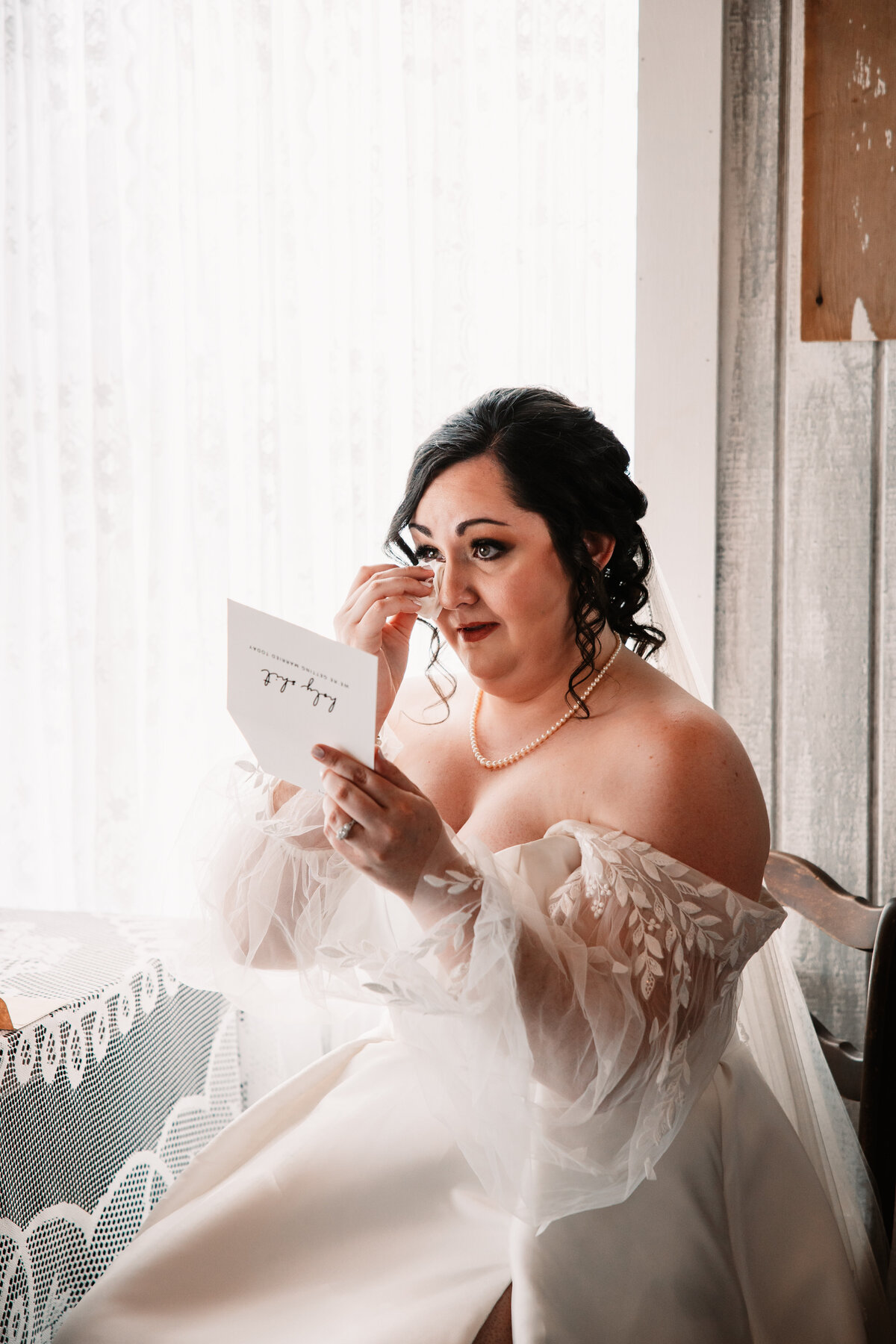 A bride sits under a window crying while reading a note from her groom