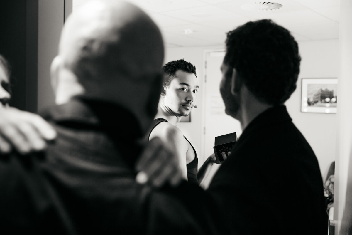 Een man in een kleedkamer kijkt tussen twee andere mannen door naar de camera.