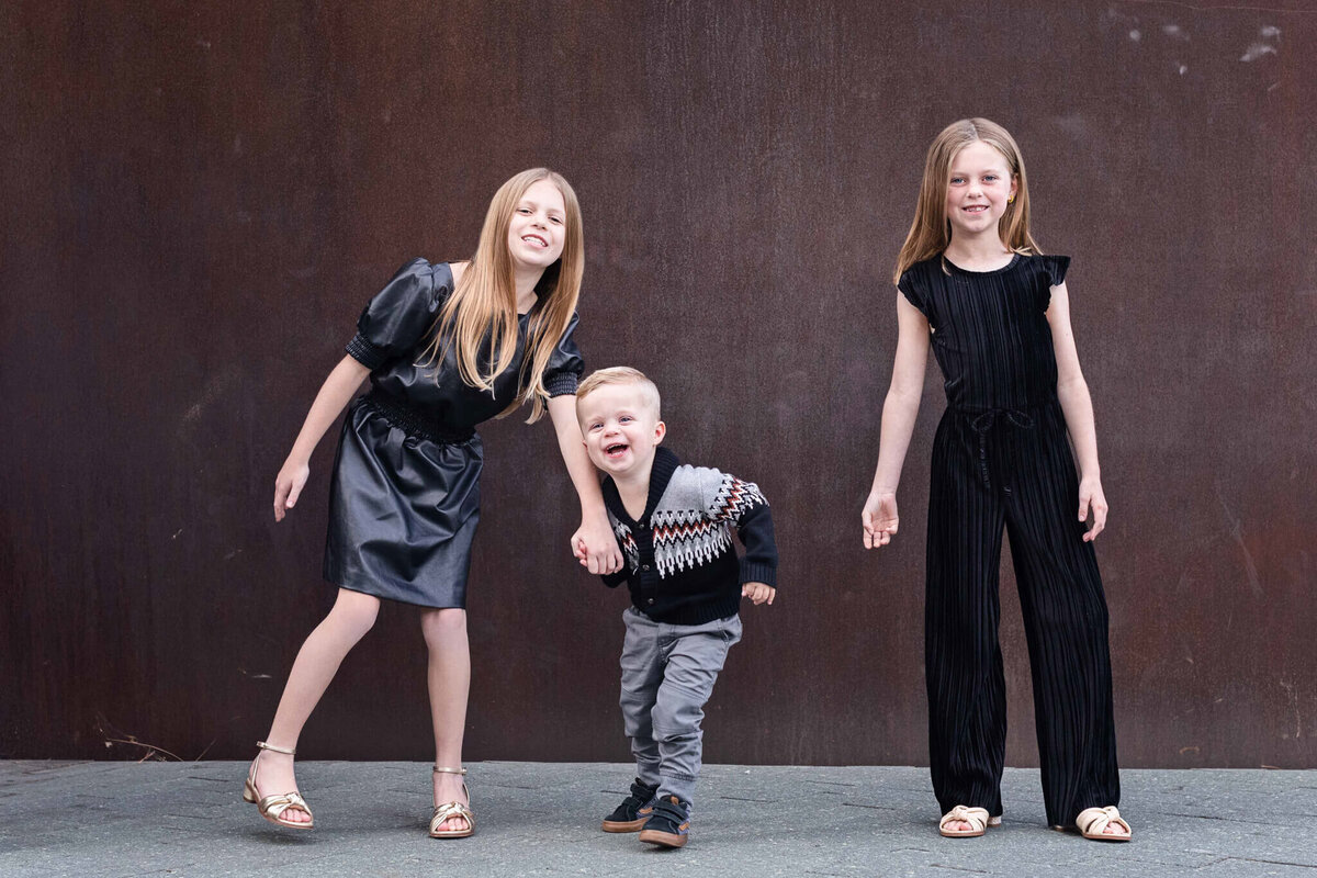 Three children (two girls and a young boy) hold hands and laugh against a brown wall at OCMA.