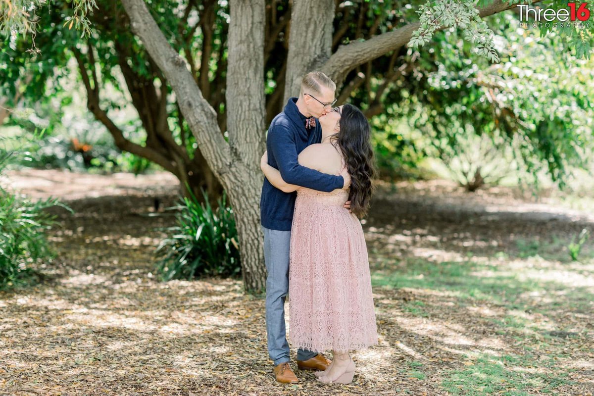 Engaged couple embrace as they kiss under a large tree