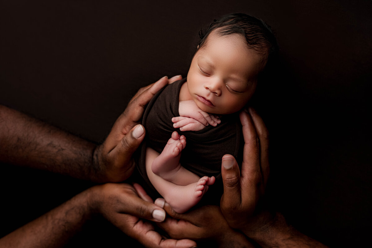 A peacefully sleeping newborn rests on a brown backdrop, cradled gently by the hands of both parents during their Charlotte newborn photography session.