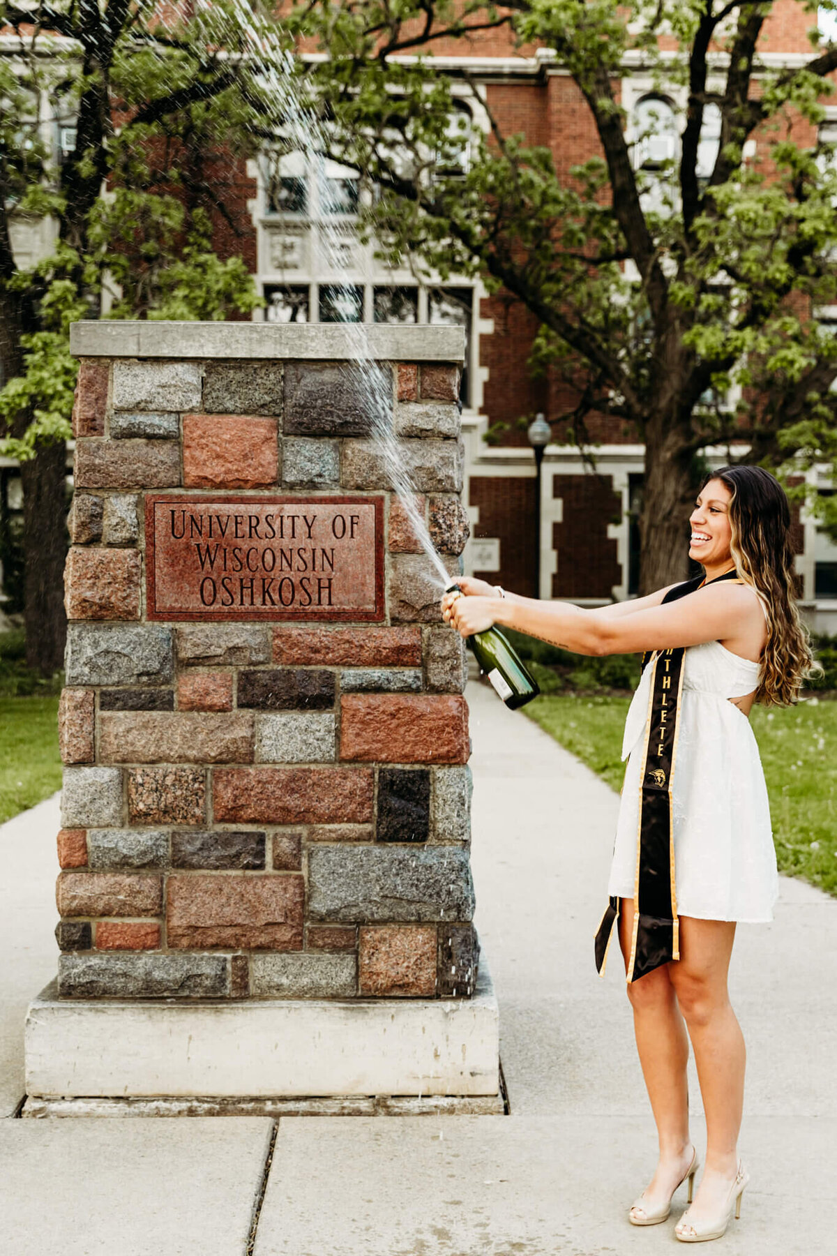 Pretty woman in a short white dress laughing as she sprays champagne in front of the UW Oshkosh sign on campus during her graduation photo session