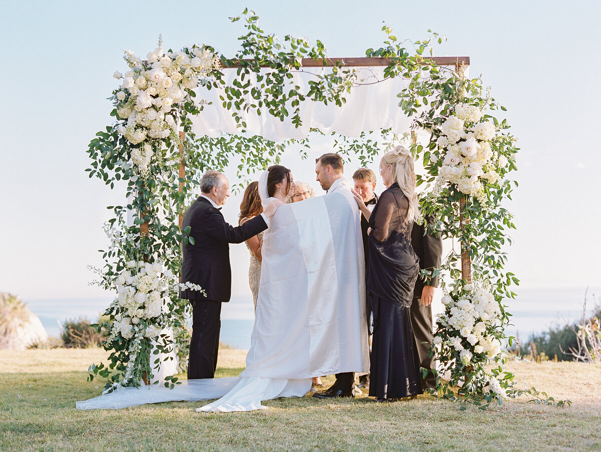 couple standing under huppah wrapped in blue and white striped blanket with white flowers