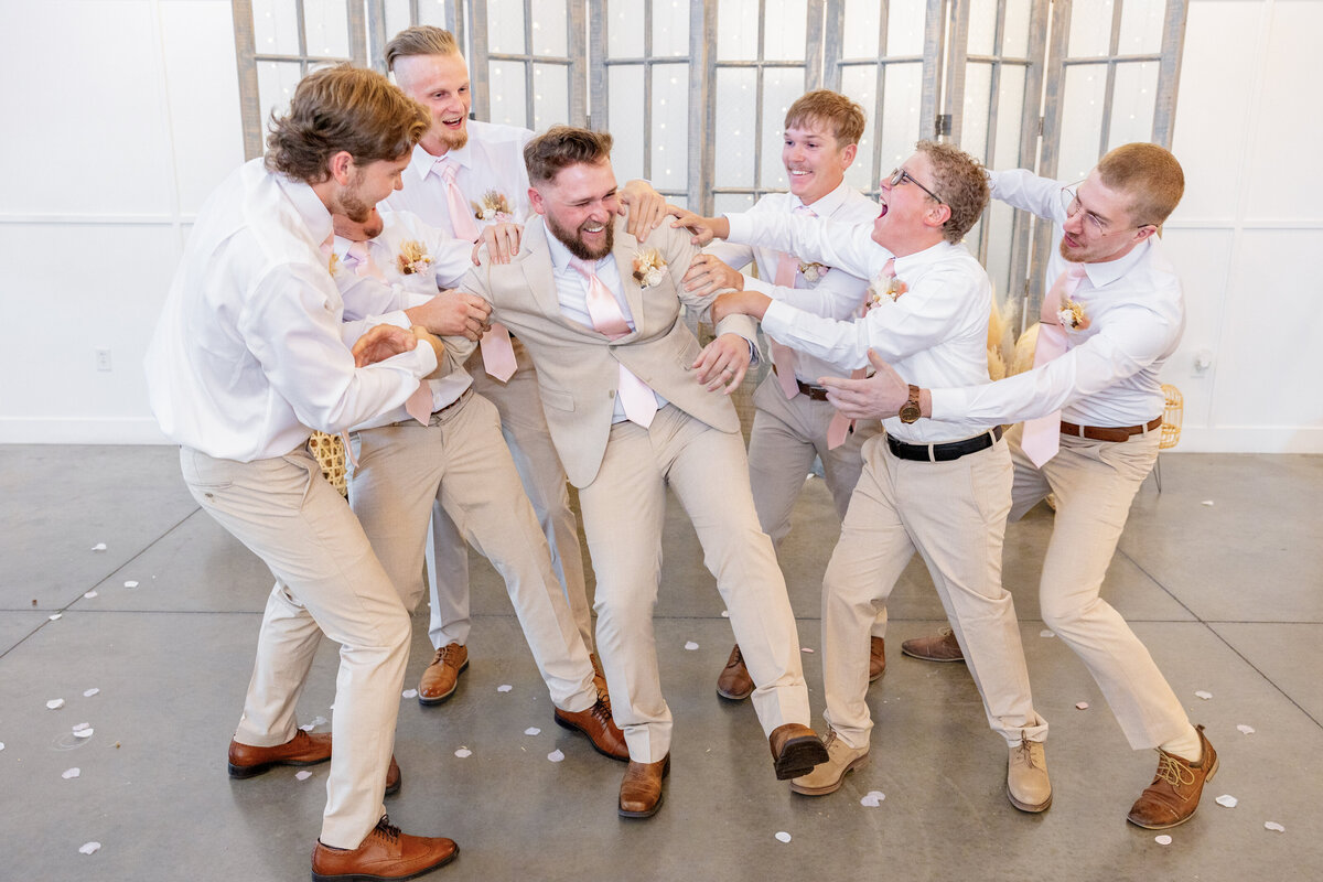 The groomsmen gather around the groom, playfully pretending to pull him away in a fun, lighthearted pose.