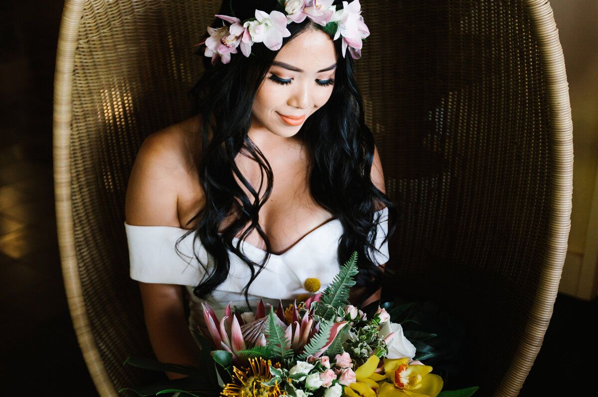 bride sits in wicker chair and looks at flower bouquet
