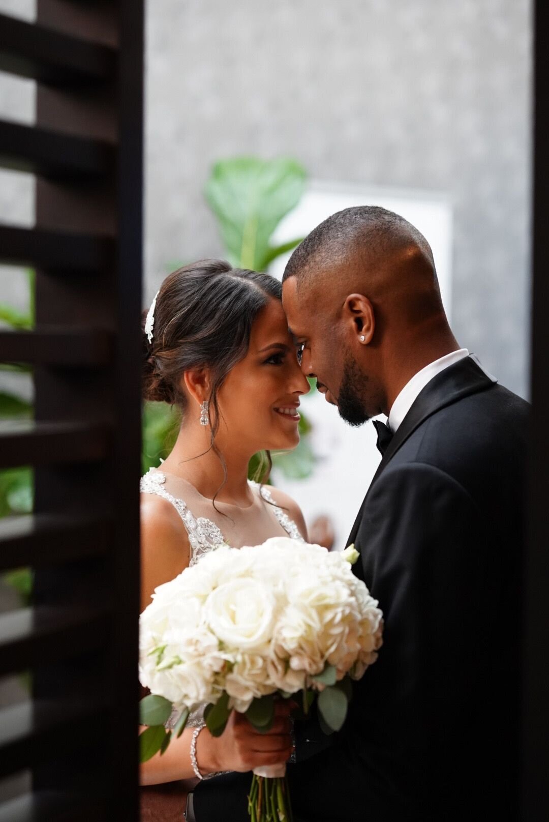 A close-up shot of a couple framed by an open door, capturing a tender moment between them. The bride and groom are seen through the doorway, their expressions full of love and intimacy. The image focuses on their connection, with the open door adding a soft, romantic framing effect.