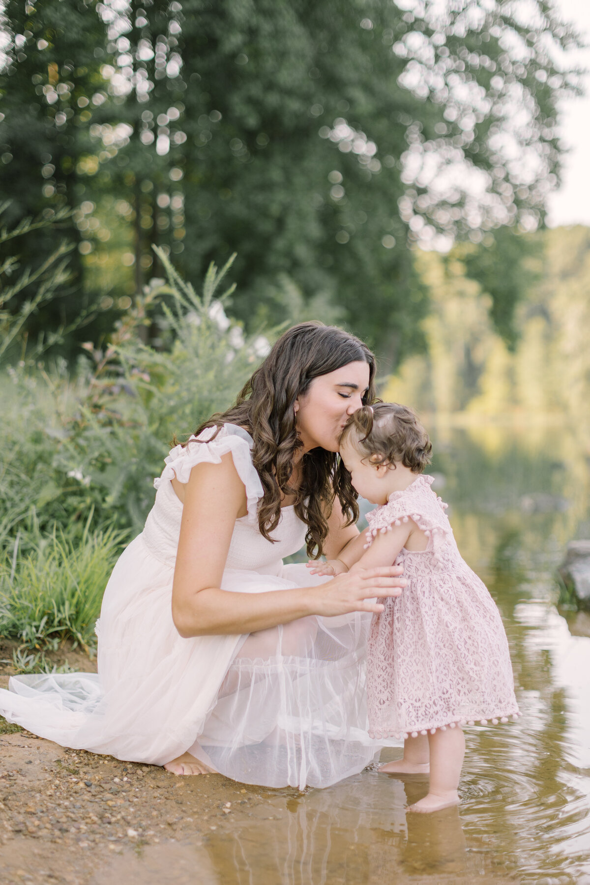 A mother kissing her toddler beside a quiet lakeside at golden hour — Portrait photographer in Raleigh.