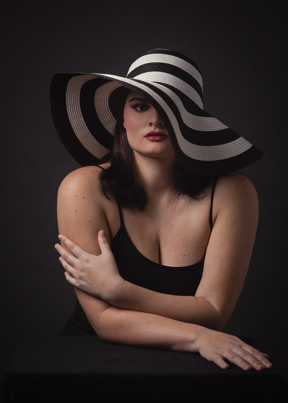 woman in black top with a black and white striped hat resting hands elegantly on a studio surface