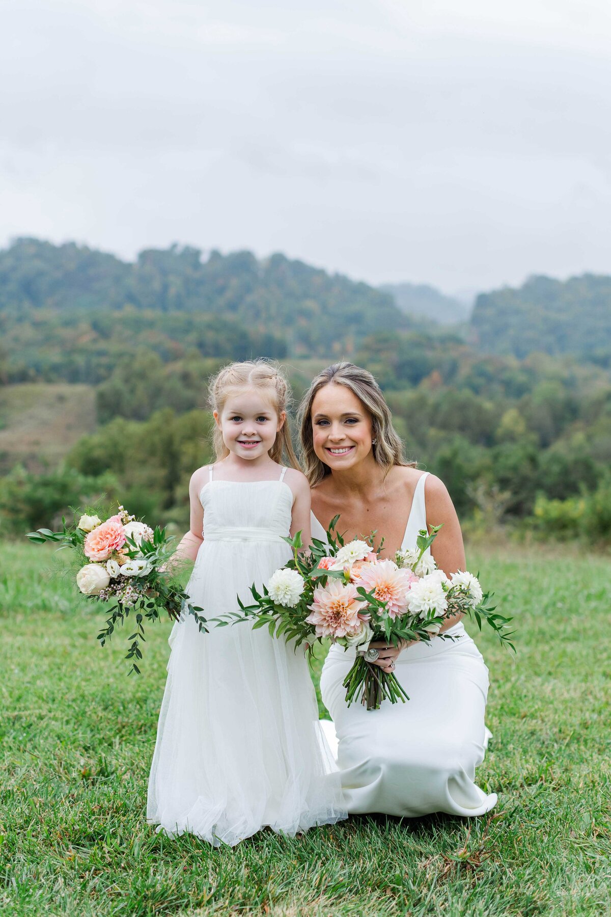 Bride kneeling beside flower girl with foggy mountains in background