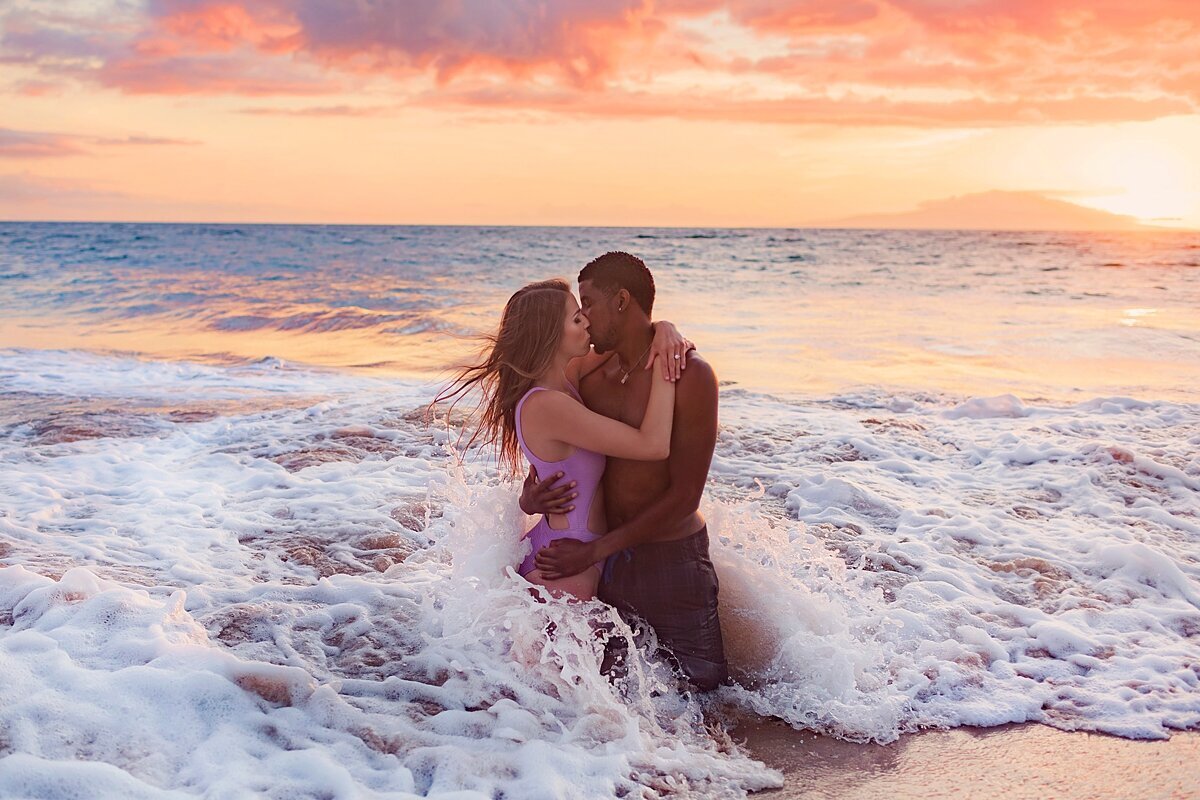 Engagement session on Maui by Love + Water featuring couple kissing while getting splashed by a wave at sunset