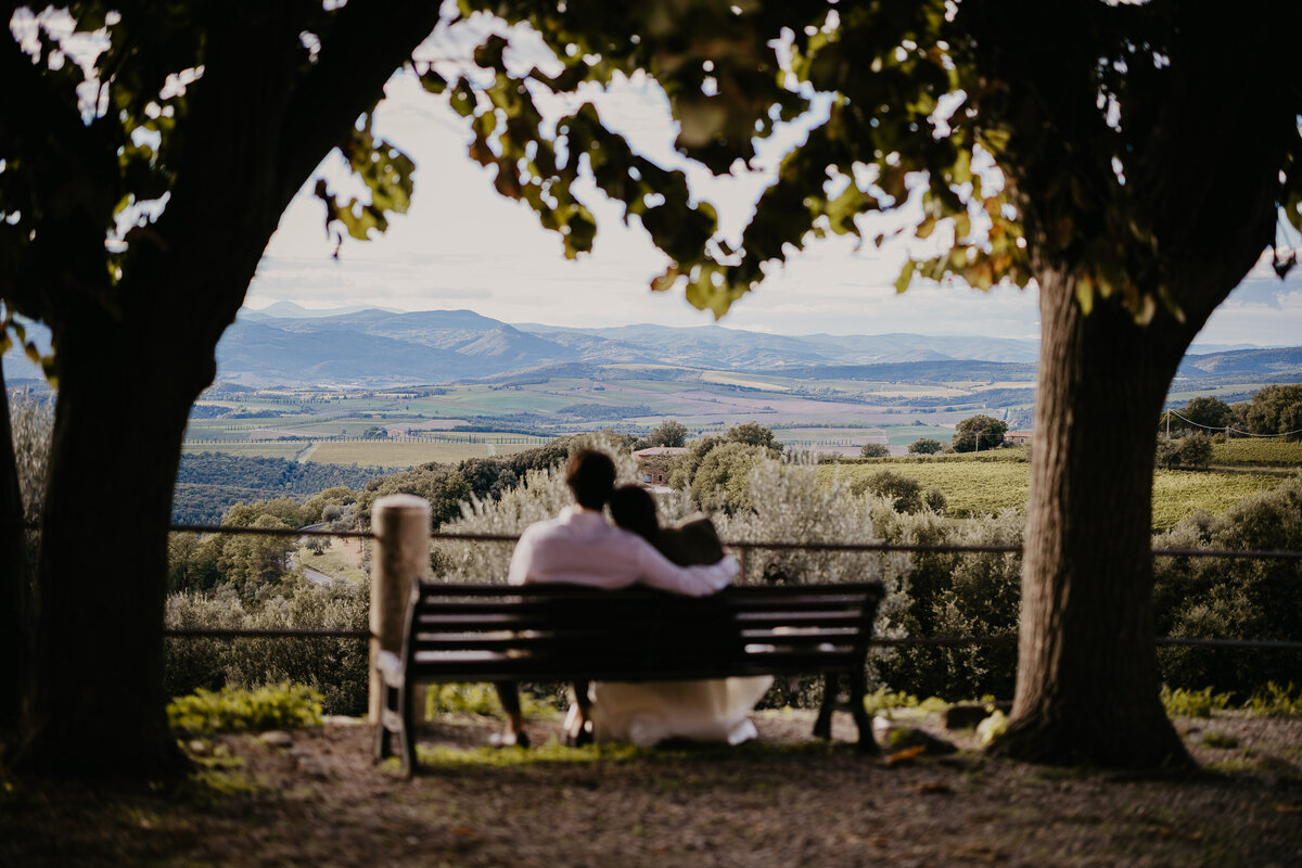 Bride and groom embracing on bench overlooking Montalcino hills, wedding photographer Tuscany.