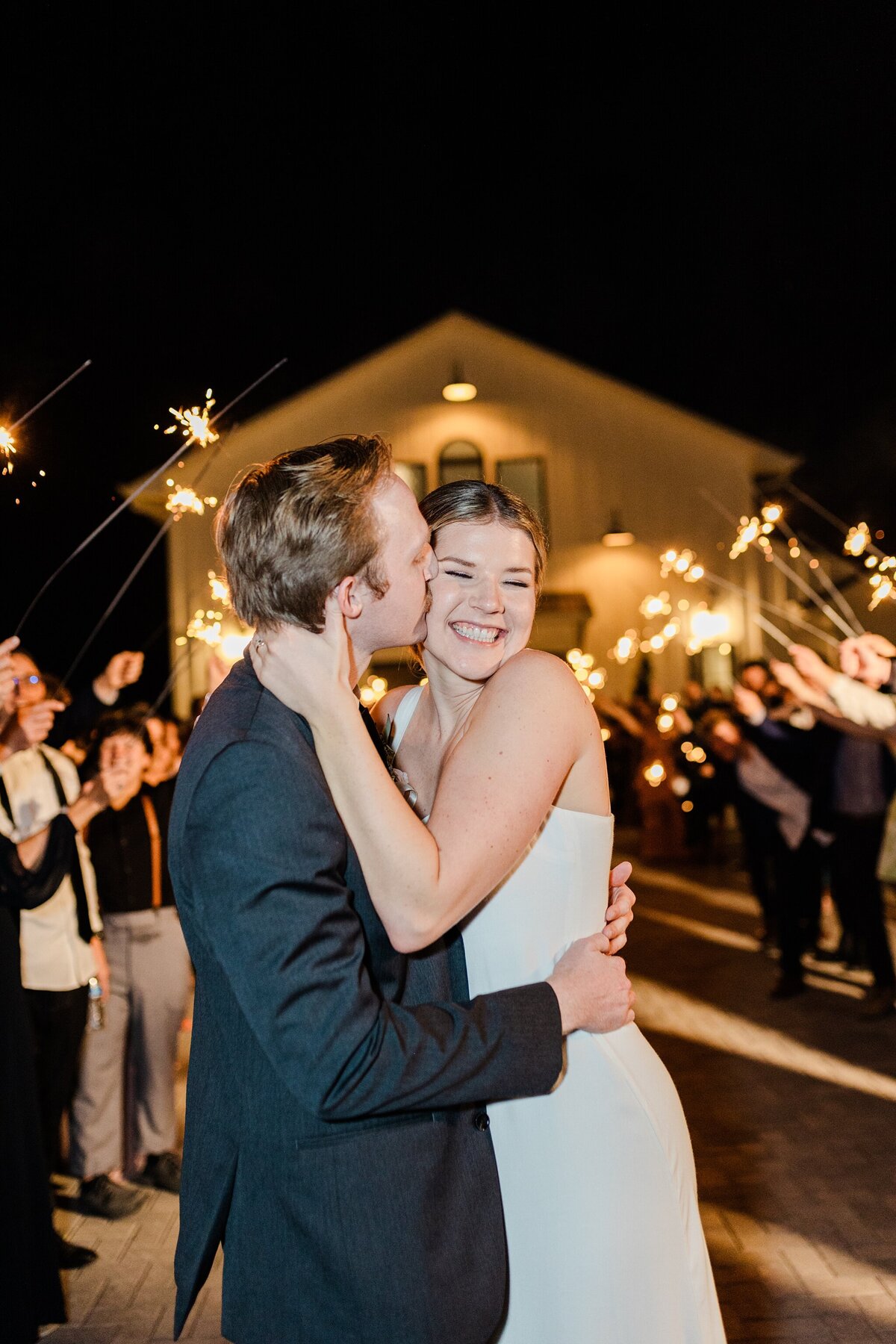 A bride receiving a kiss on the cheek from a groom during their sparkler exit after their wedding reception in Dallas, Texas. The bride is on the right and is wearing an elegant, sleeveless, white dress. The groom is on the left and is wearing a dark suit. Their guests create two long lines on either side of them and hold up lit sparklers in celebration.