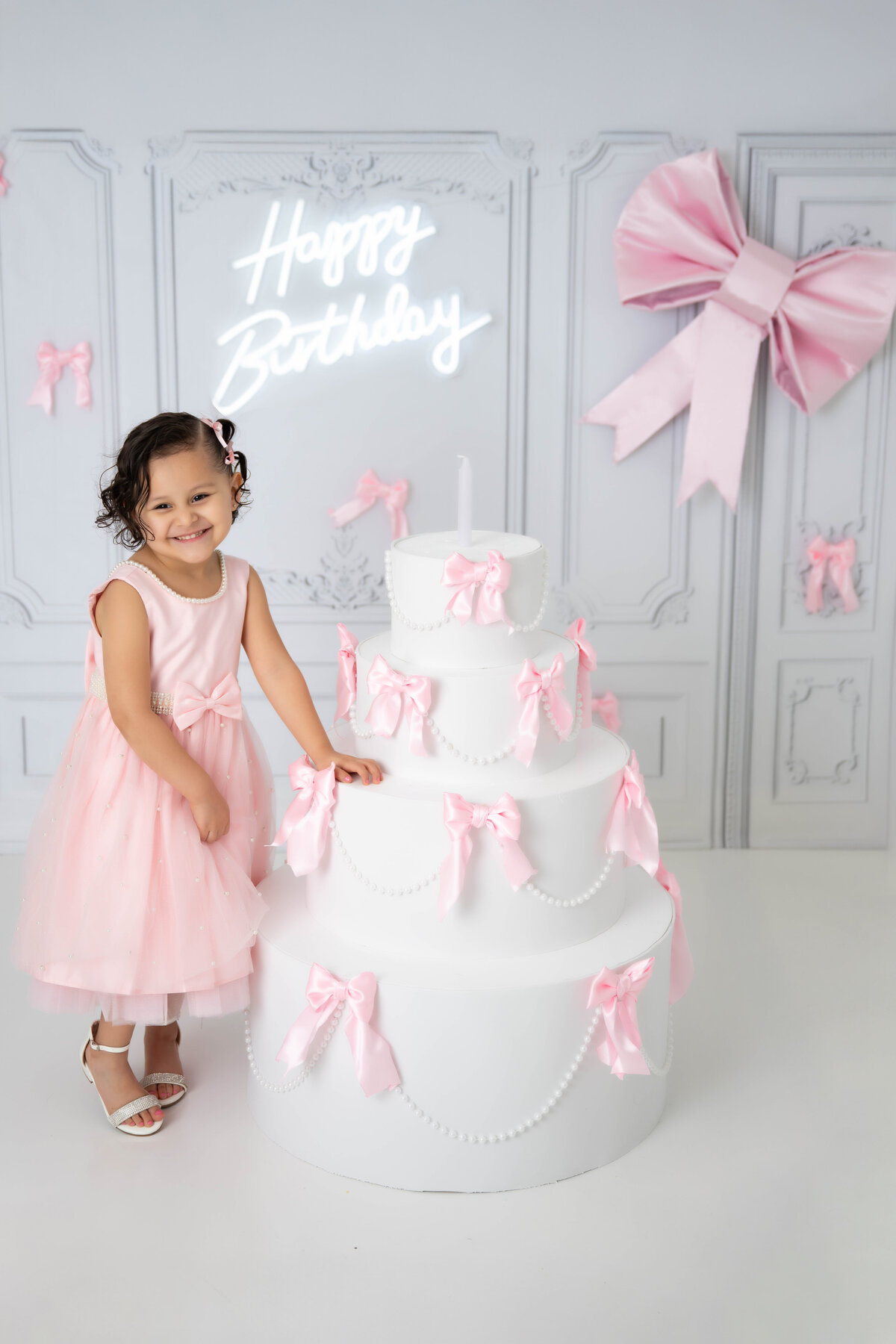 Toddler standing next to a large cake prop with pink bows and a "Happy Birthday" sign in the background.