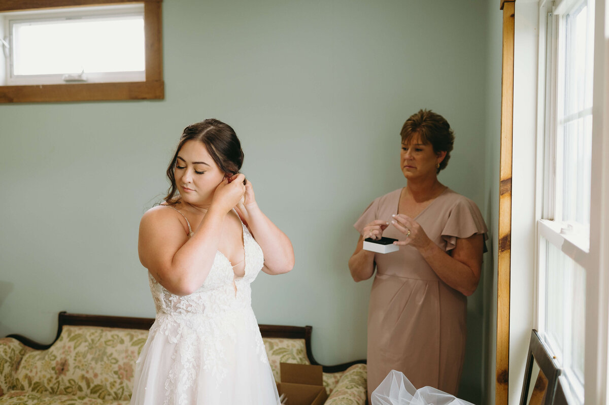 mother of the bride watches as her daughter puts on her earrings in a bridal suite that is light blue with wood furniture captured by Charlottesville wedding photographer