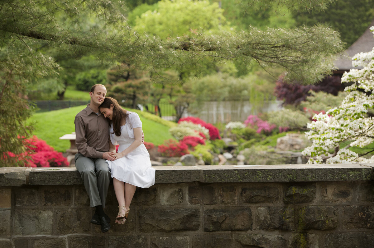A couple sitting on a small stone wall leaning into each other 