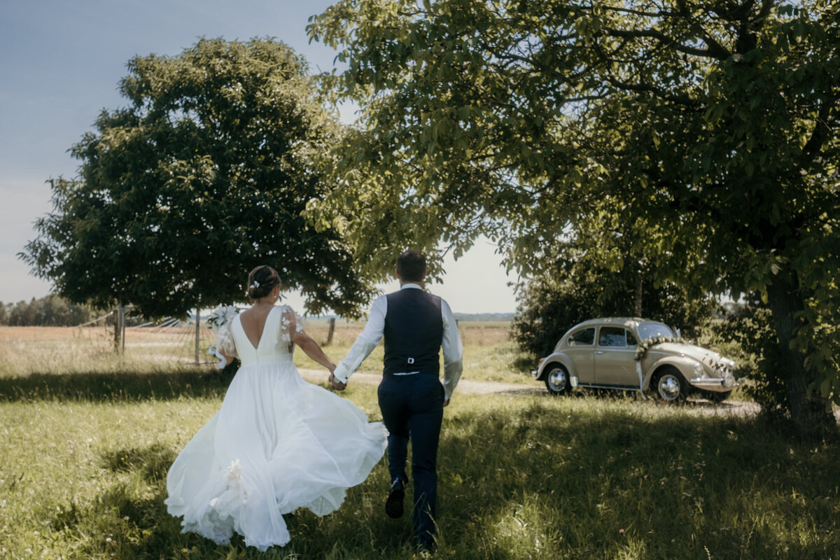 Das Hochzeitspaar rennt über eine Wiese auf den Hochzeitsoldtimer zu. Ihr Kleid weht im Wind.
