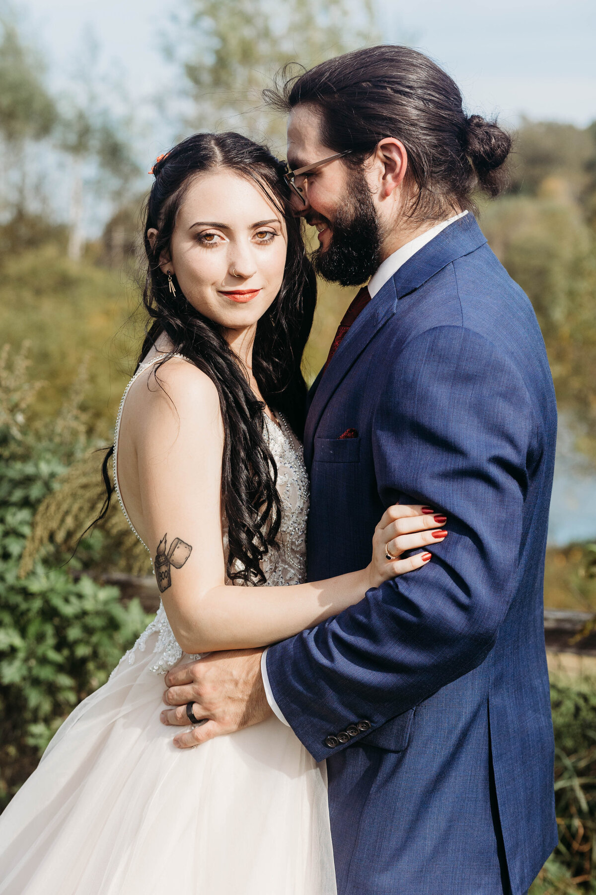 A groom in a blue suit nuzzles his bride's temple as she smiles and stands in a garden