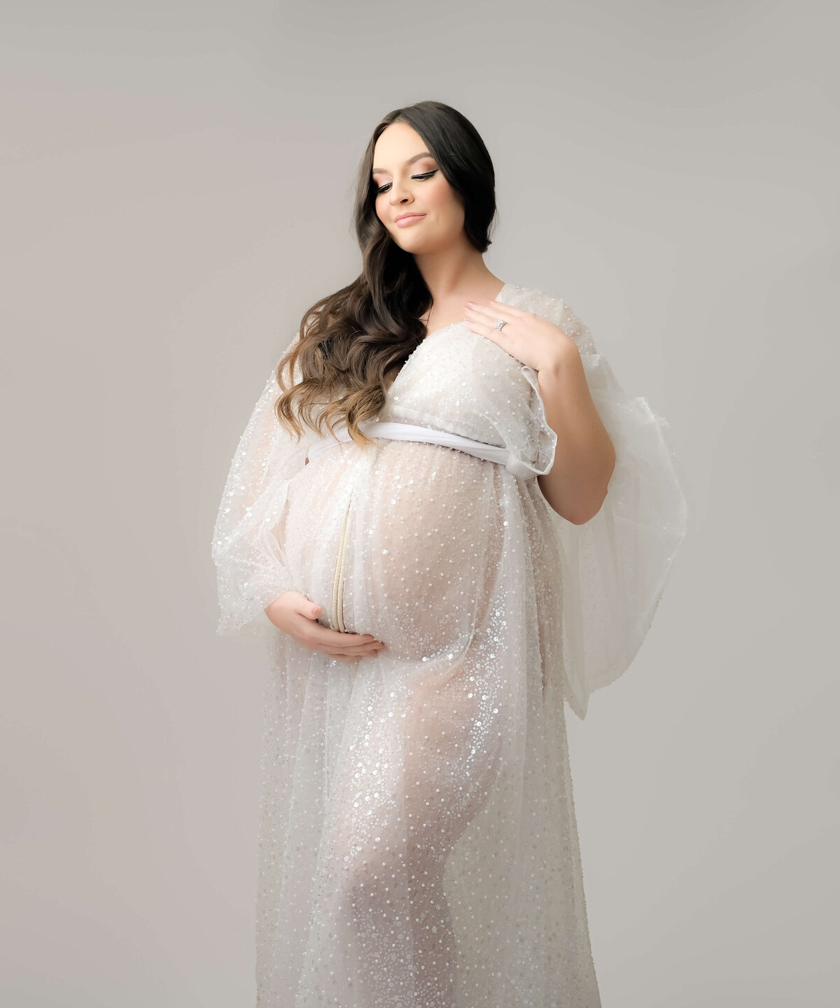 This maternity photo session takes place in Bremerton. The expecting mother is in a white sequin sheer robe. Photographed from a slightly low angle looking up, her long brown hair rests on one shoulder  as she looks down to her belly.