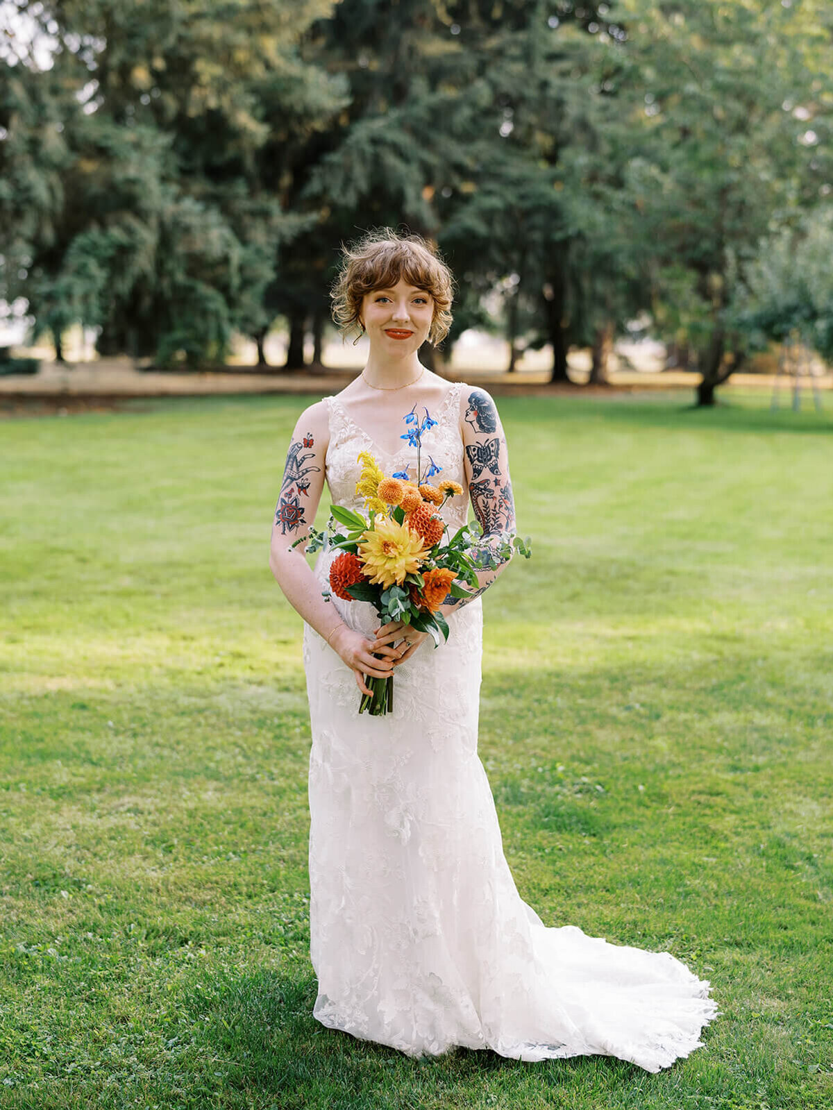 A bride with tattooed arms smiles while holding her colorful bouquet. 