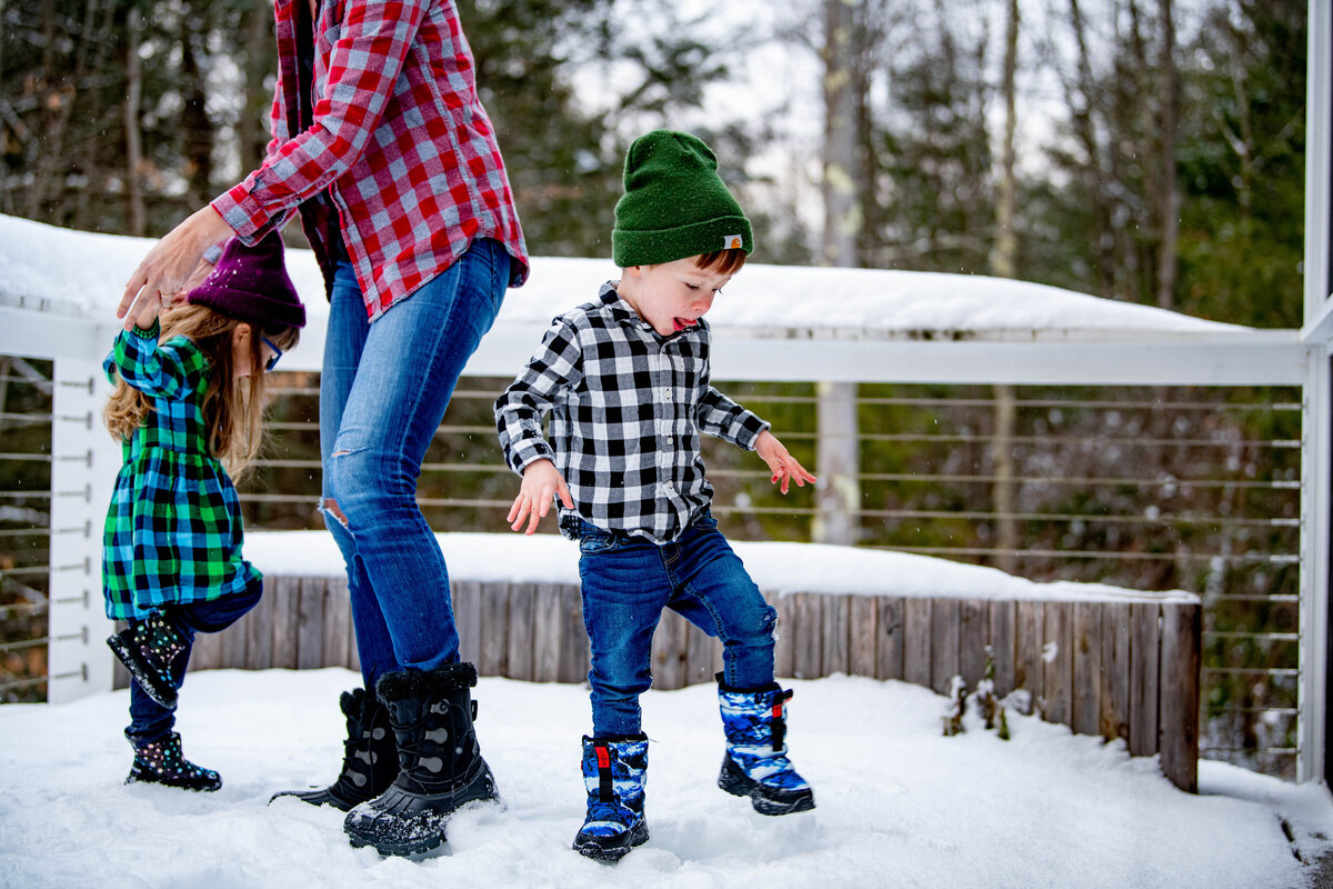 a little boy wearing boots dances around his mother and sister in the snow