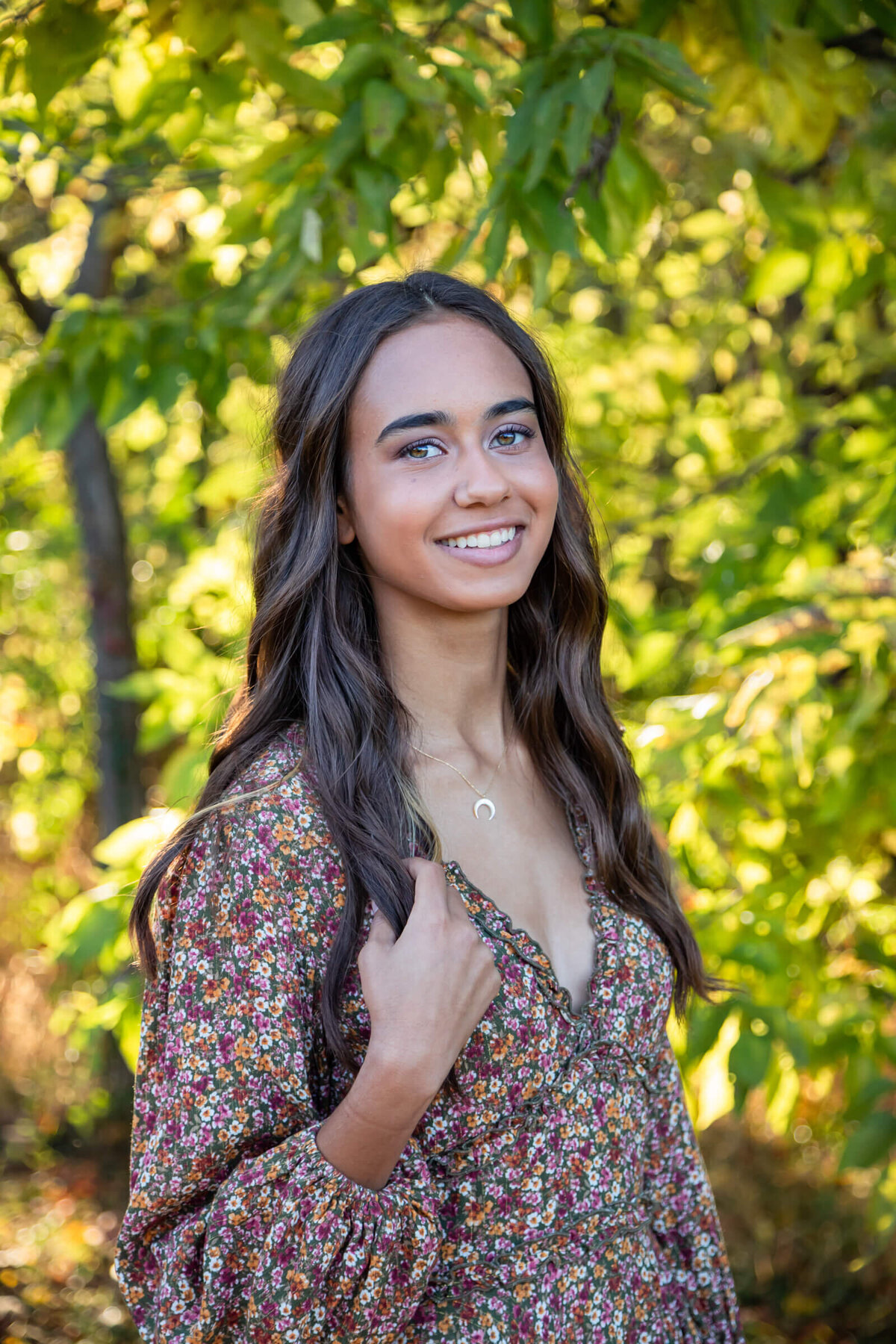 A stunning dark haired teenage girl poses for a senior picture in a brown floral dress surrounded by green leaves. Captured by senior photographer Dynae Levingston.