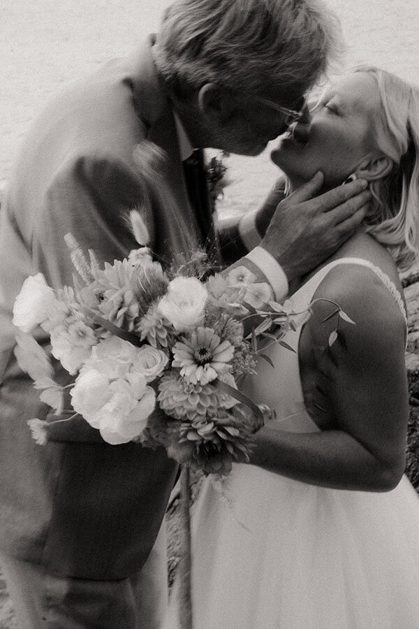 Le marié embrasse tendrement la mariée dans une douce étreinte, bouquet en main. Photographie en noir et blanc lors d'un mariage en Bretagne par Kamescop, photographe et vidéaste.