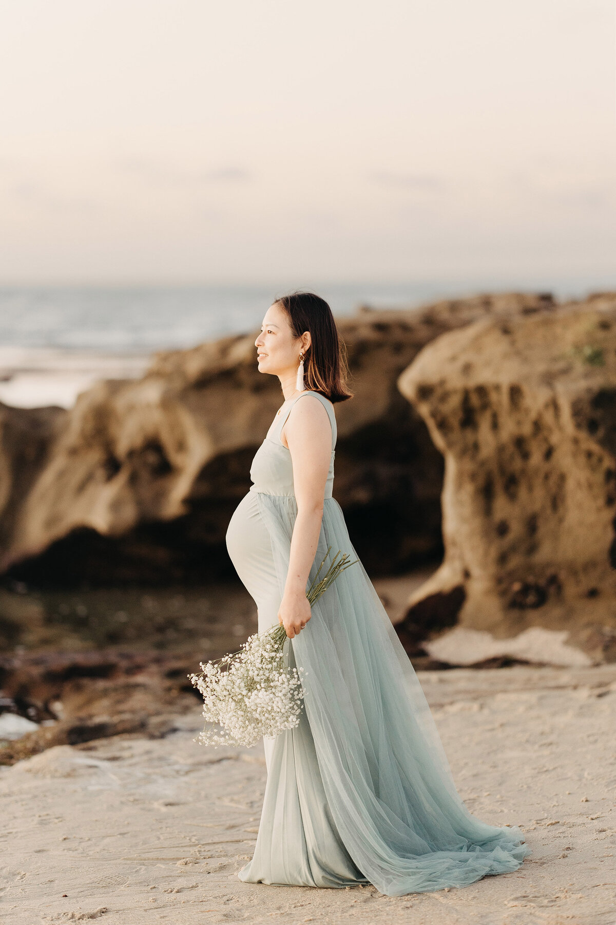 Expecting mother in a flowing embroidered gown during a maternity session on the rocky coastline of La Jolla at golden hour. Captured by San Diego Family Photographer Yuka Schad.