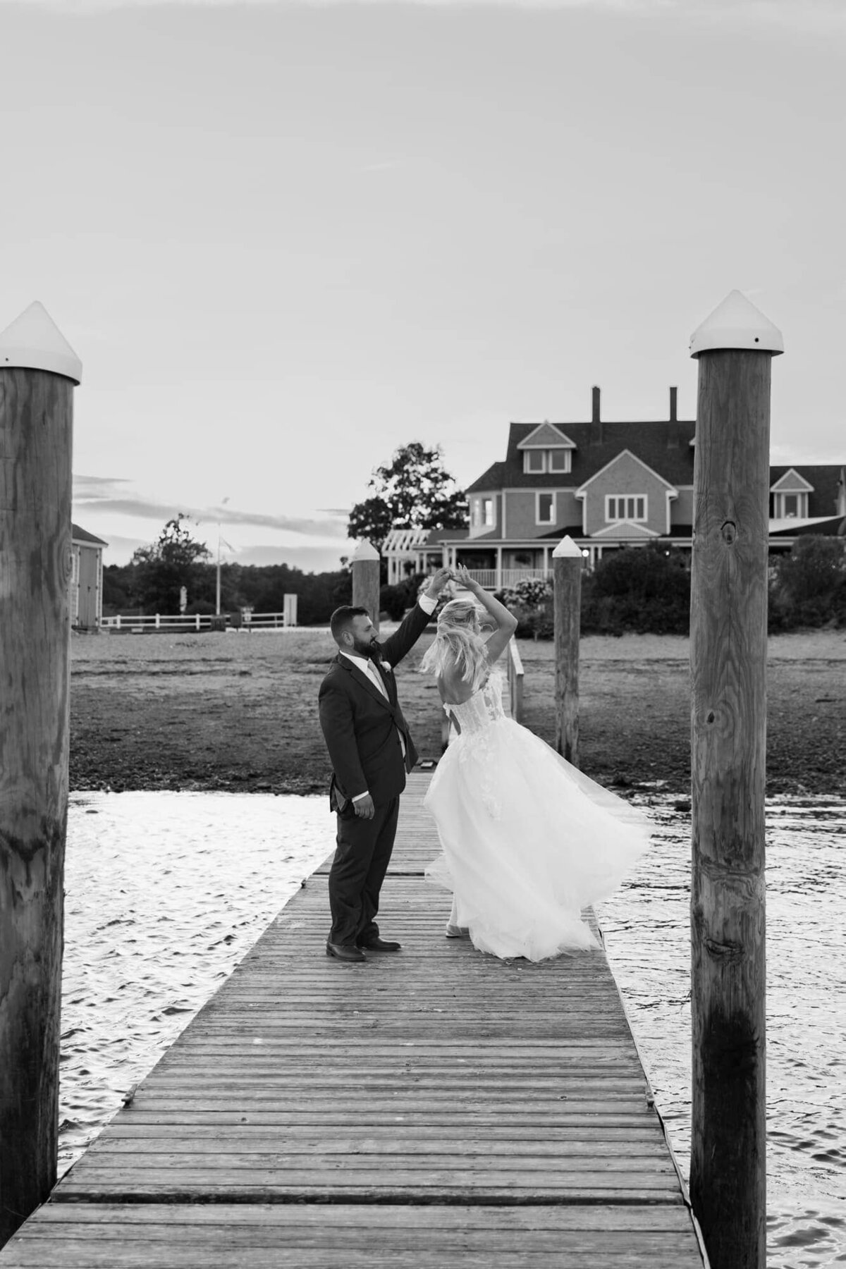 Rhode Island Wedding Photographers | Kelsey Sheehan Photography Timeless Rhode Island Weddings | A bride in a flowing gown and groom in a suit dance on a dock over calm water, with a large house and trees in the background, conveying joy.