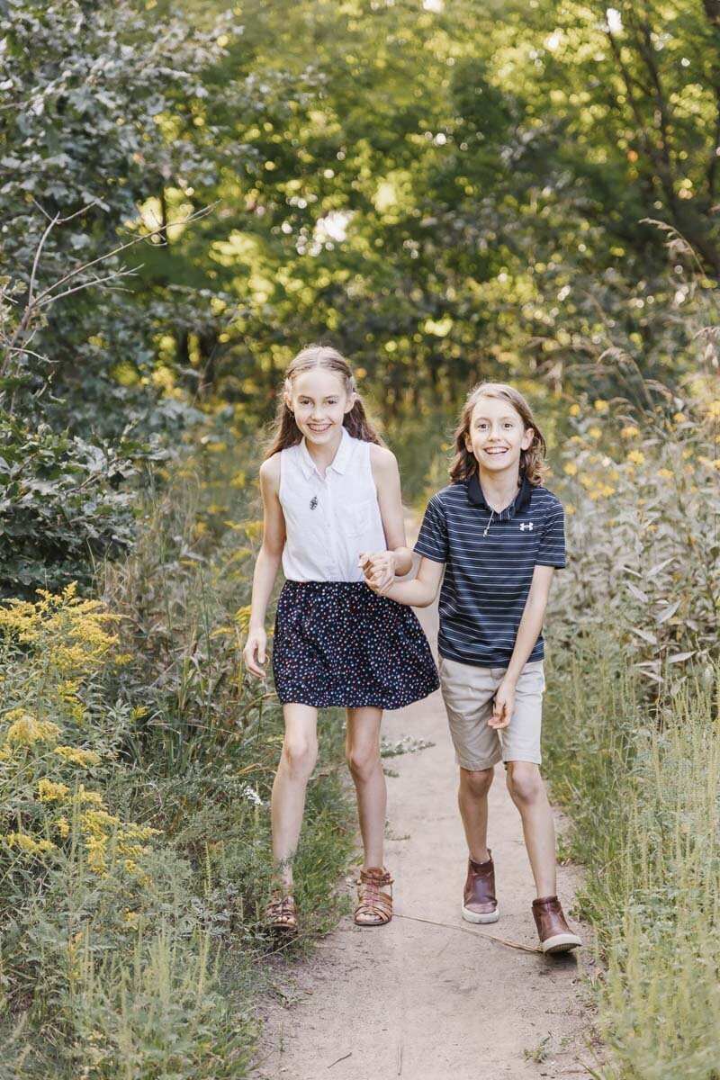 Two children walk hand in hand on a dirt path surrounded by greenery and yellow flowers, capturing the essence of summer family portraits at High Park Toronto.