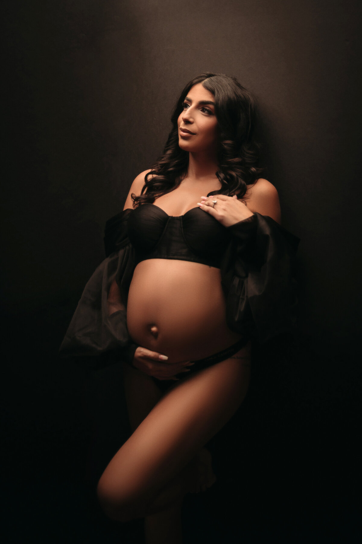 brunette pregnant woman wearing black lingerie holding her baby bump in front of dark backdrop