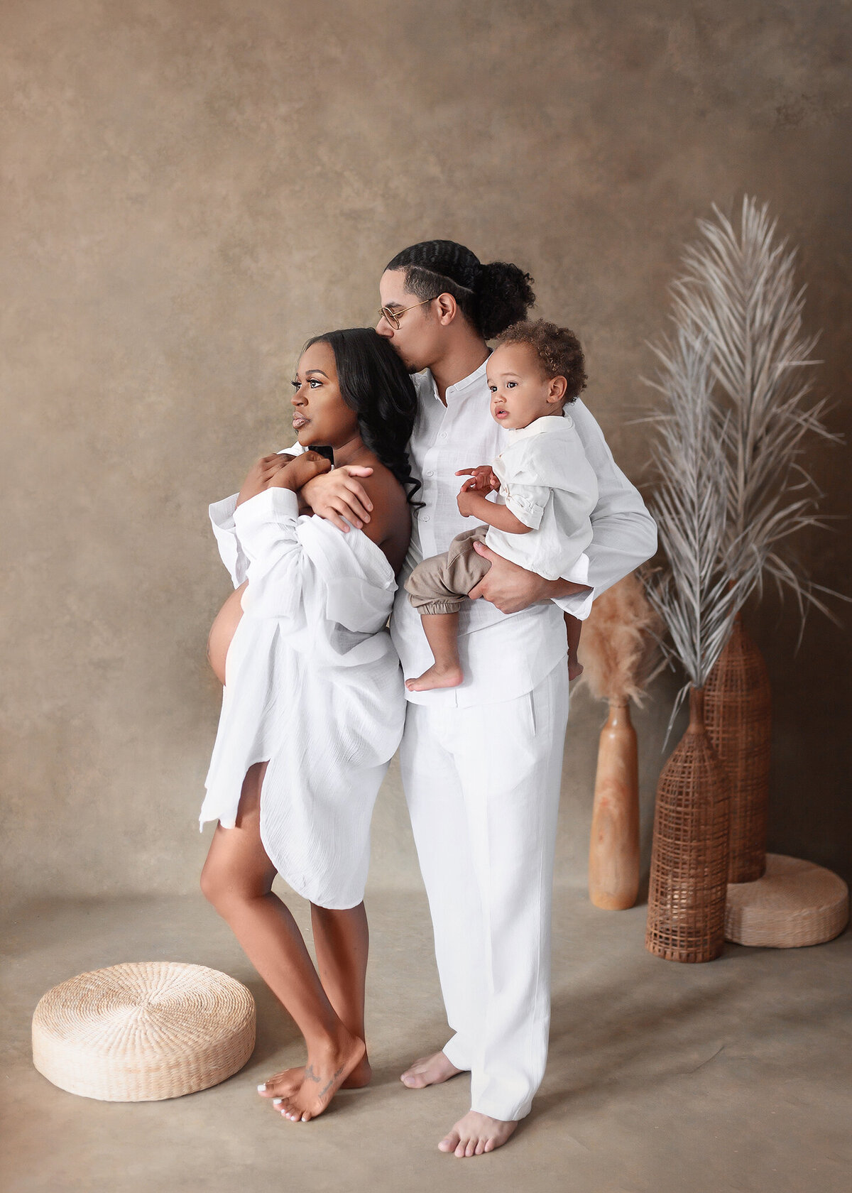 Expecting couple along with toddler chilld on a light brown background wiht tropical  feeling background with dried palm leaves, rattan , wearing white