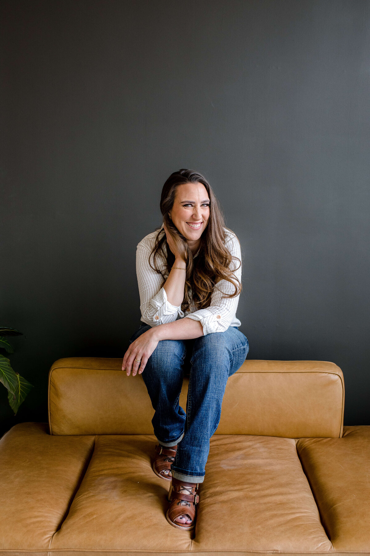 commercial photographers captures brand photography for small business owner sitting on a brown leather couch posing as she sits on the back of the couch with a black wall behind her