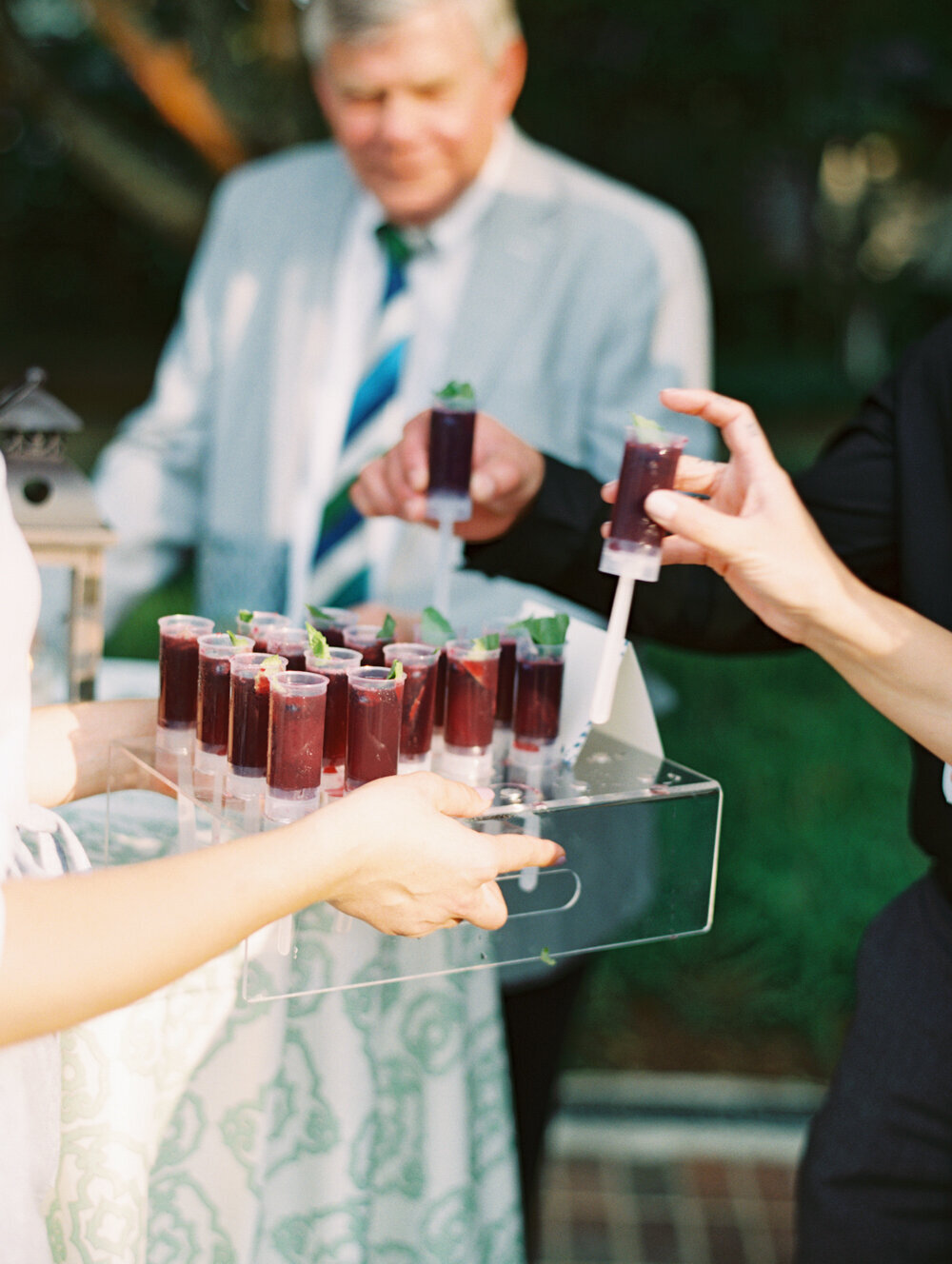 At The Hamptons, New York, waterfront estate tented wedding captured beautifully by photographer Bonnie Sen and videographers East West Films, we see a person holding a clear tray filled with purple liquid-filled push-up popsicles, each adorned with a delicate mint leaf. Guests are eagerly reaching out to grab the delightful treats. In the background, a person dressed in a light suit and striped tie adds a touch of elegance, though slightly out of focus, creating a charming ambiance to the scene.