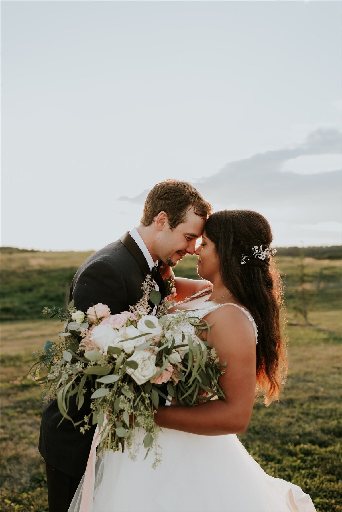 Bride and groom standing forehead to forehead, bride holding a bouquet with sunset behind them.