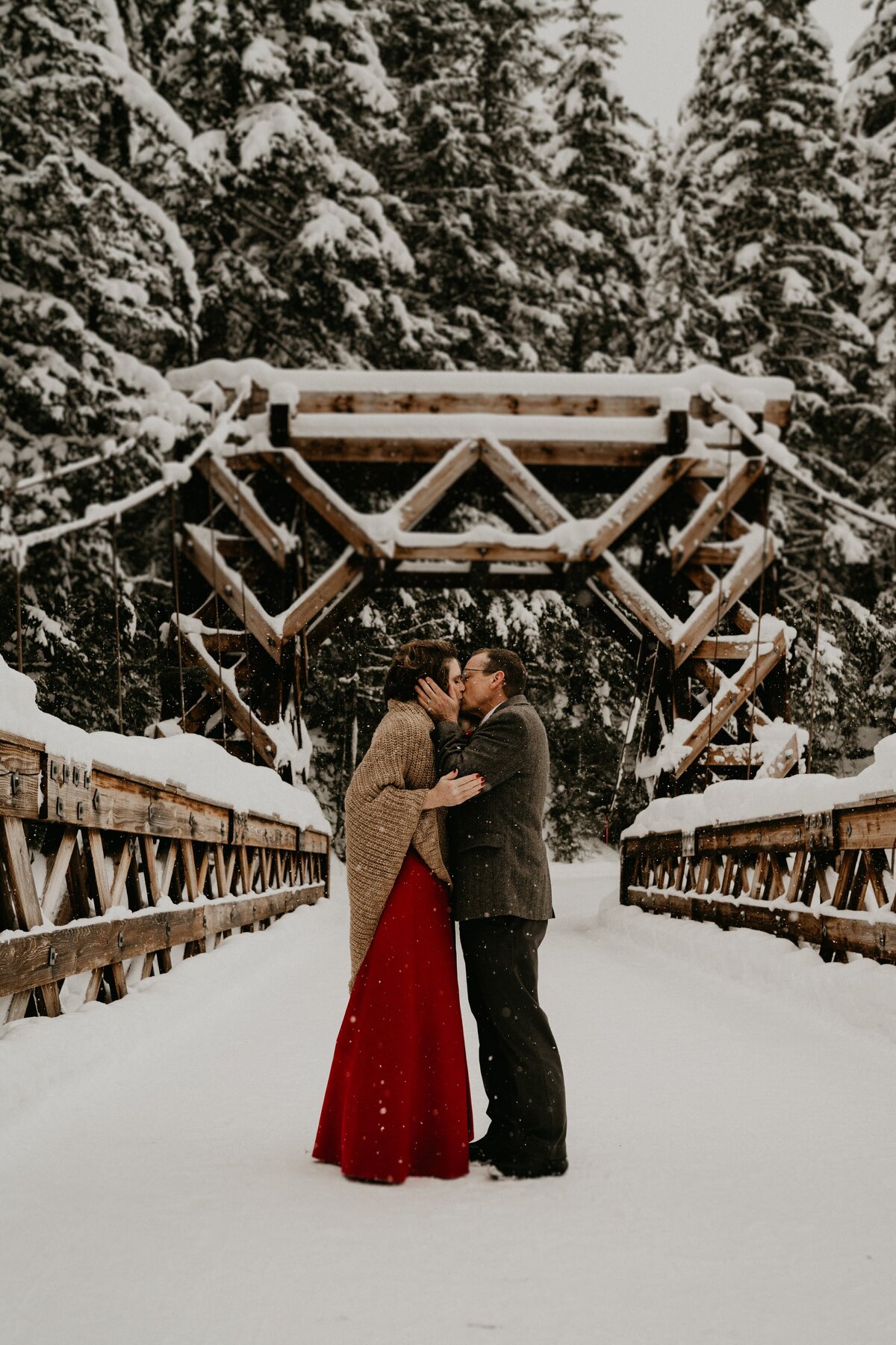 Bride in red dress & groom kiss on snowy historic bridge during their winter elopement at Mount Rainier National Park!