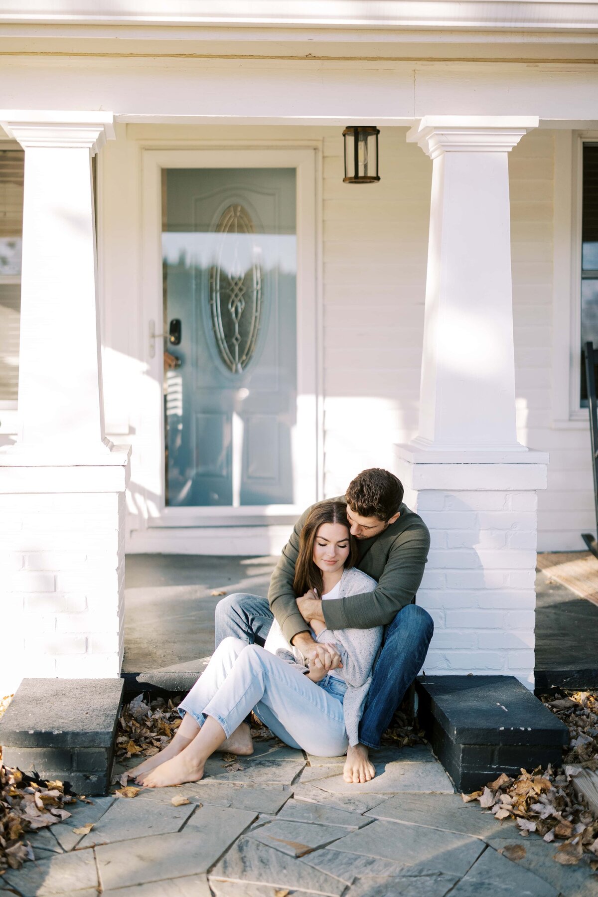 Young couple in comfortable clothes cuddling up together on the front porch of a white house with a pale blue door.