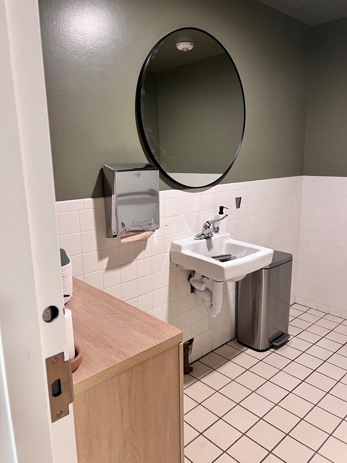 Minimalist restroom with white tile wainscoting, olive-green upper walls, round black mirror, and a wall-mounted sink.