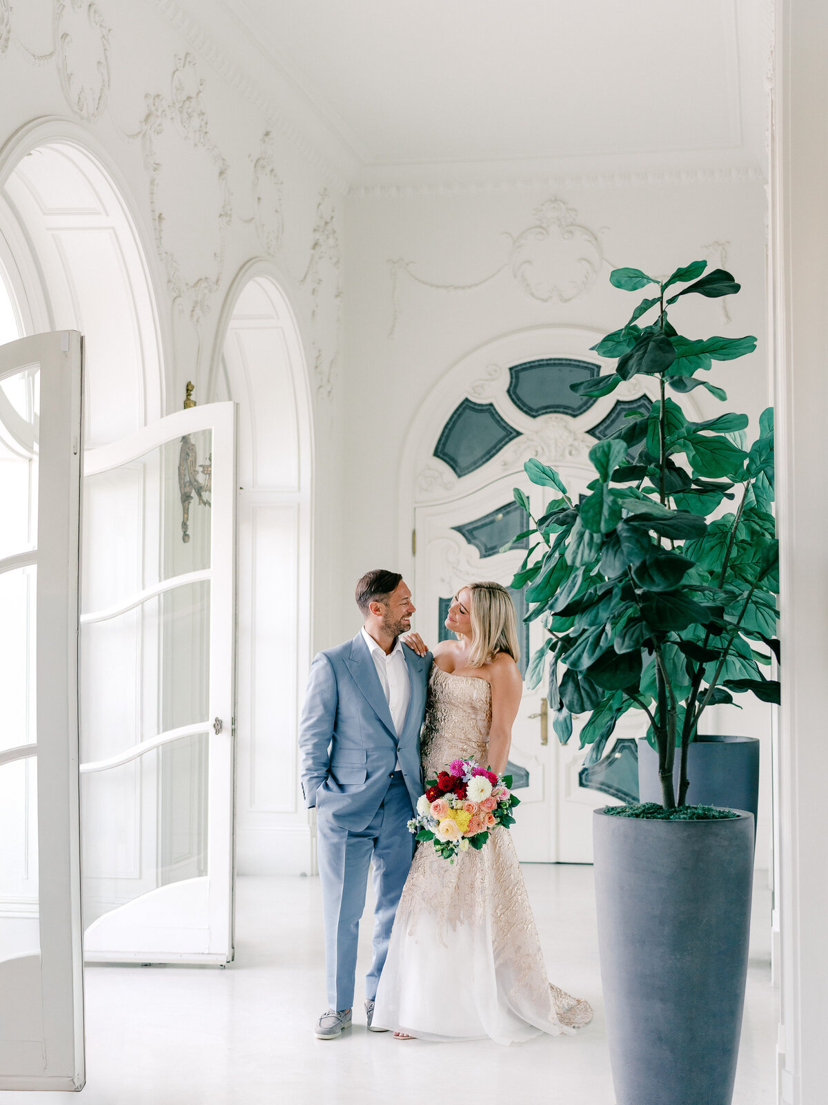 Bride in gold dress and groom in light blue suit looking at each other in a bright white private estate hallway in California 