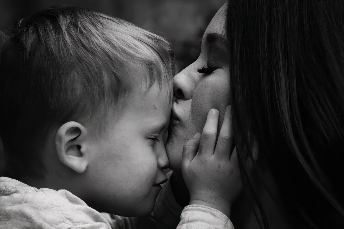 close up of  white mothers face with sons face and hands embracing her cheeks as she kisses his forehead