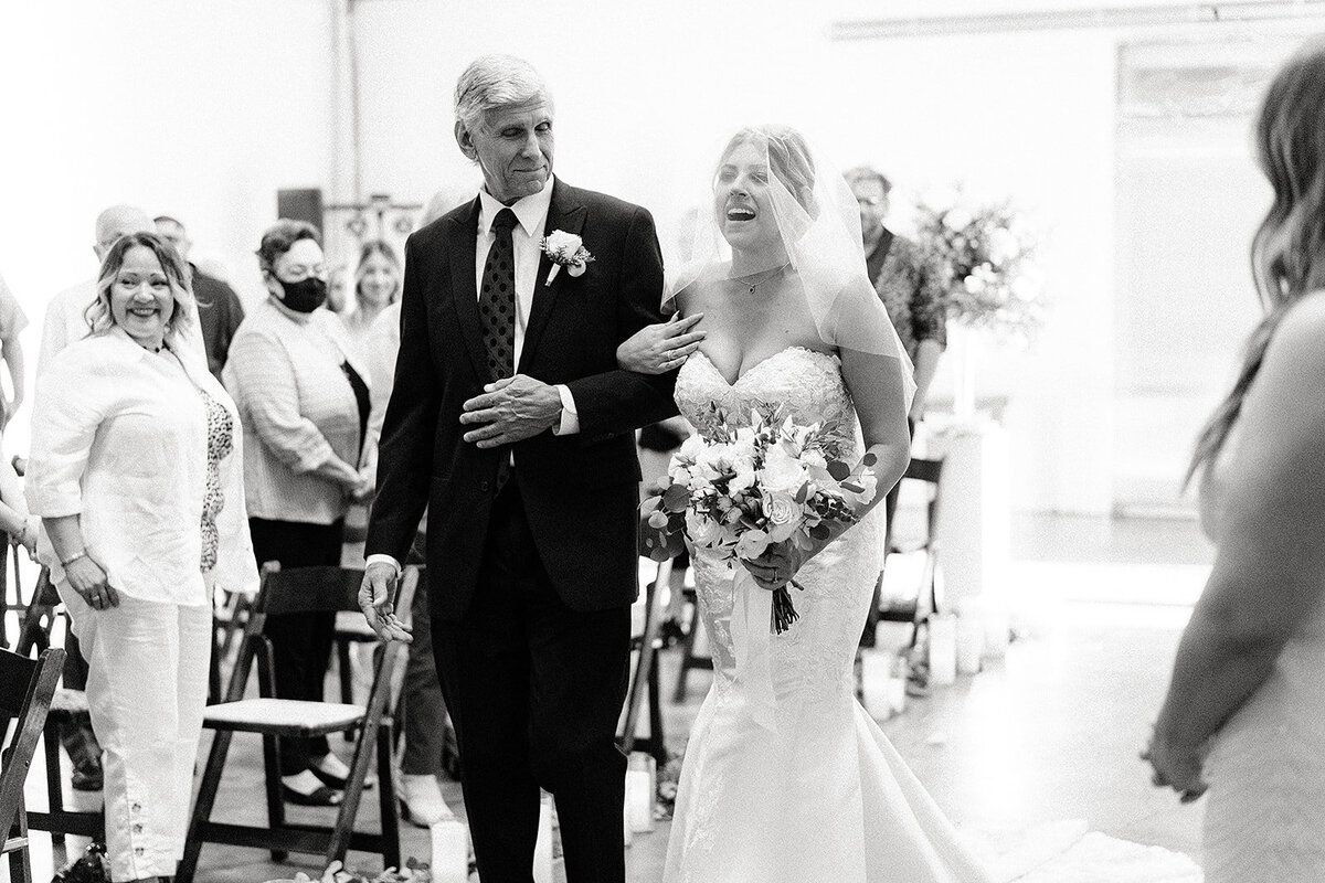 The father of the bride, smiling proudly, walks her down the aisle as she beams, holding a bouquet of white flowers and greenery, with guests looking on.
