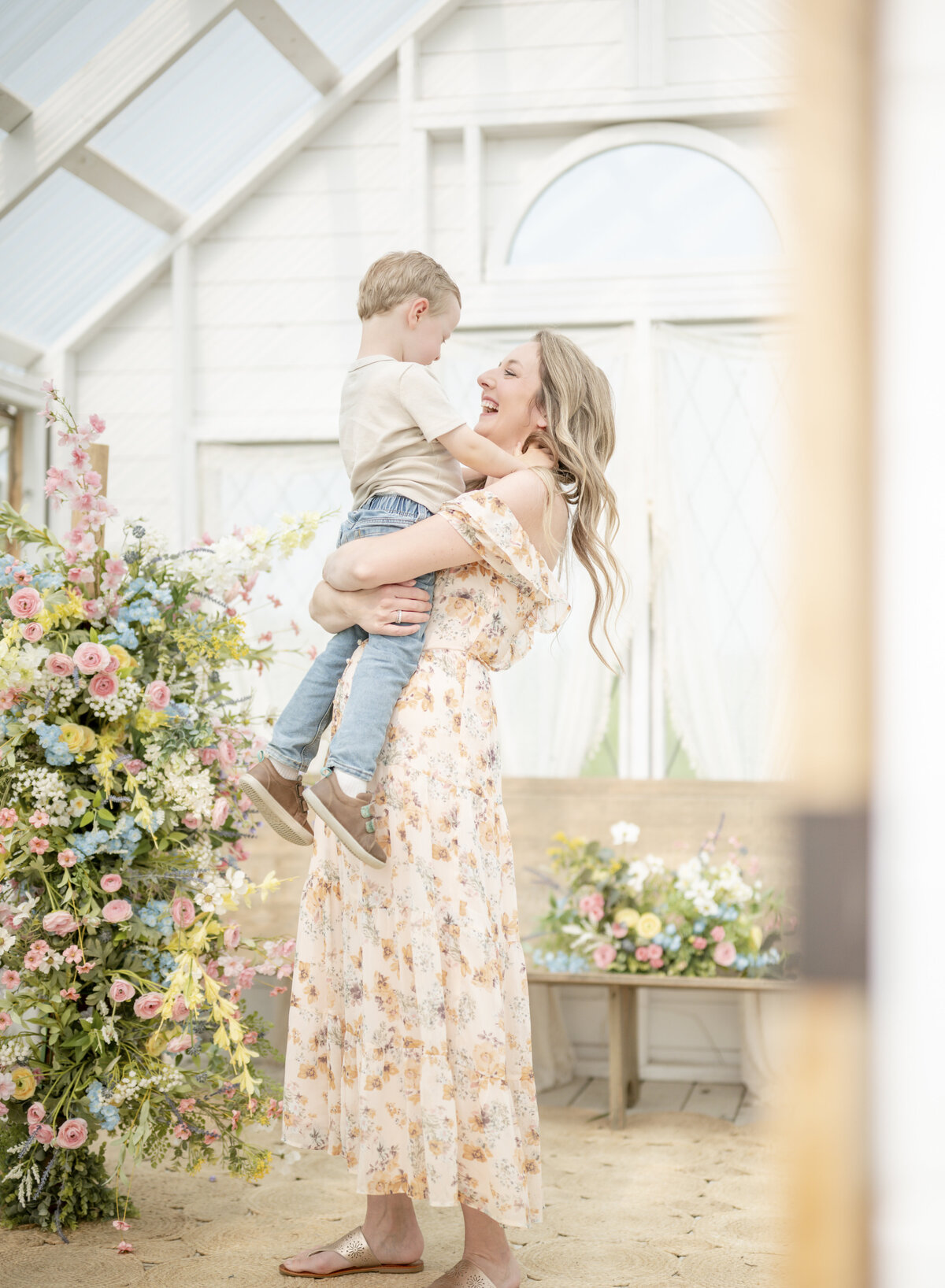 Mother lifting toddler in a floral dress in a flower-filled greenhouse during a Kansas City motherhood session.