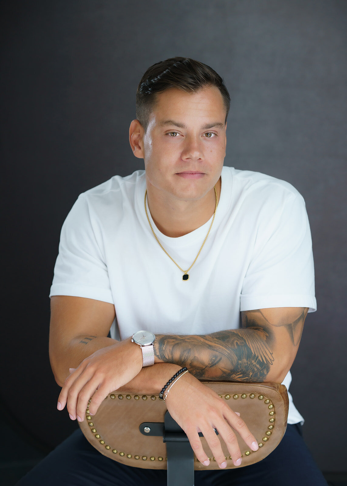 Young man with dark hair sitting on a brown chair in a white teeshirt posing for a modeling portfolio headshot