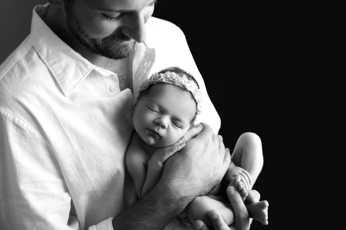 Best New Jersey Newborn photographer, Katie Marshall, captures a fine art newborn photo. In the photo, dad is holding his newborn baby girl. Baby girl is facing outward toward the camera. Baby is sleeping and wearing a floral headband. Baby's hands are folded atop of dad's. Dad is looking down and smiling at his baby.