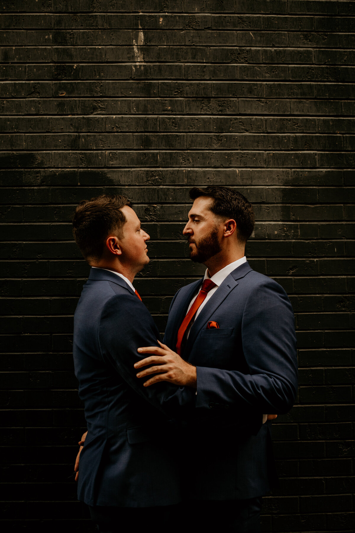 two grooms in blue suits hodling each other in front of a black brick wall in Albuquerque