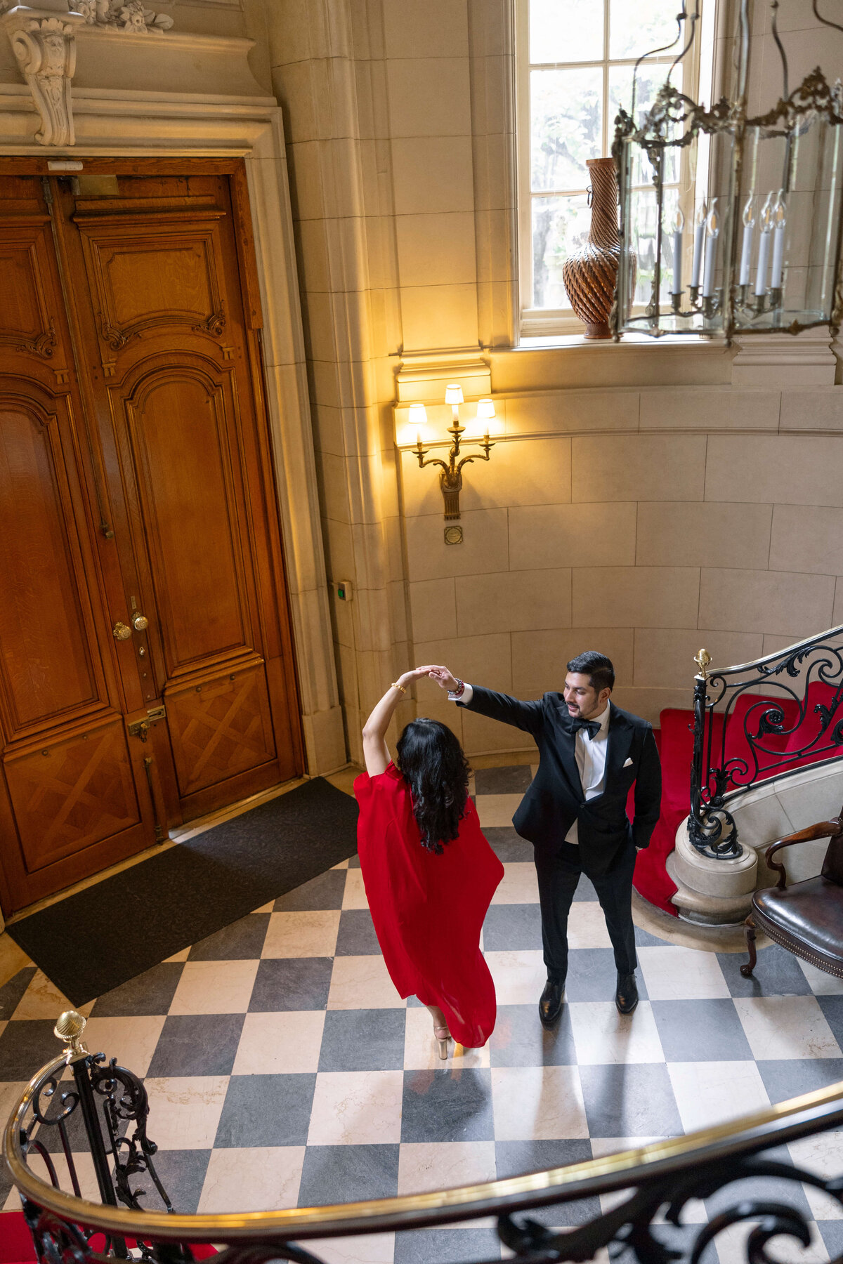 A couple elegantly dances on a checkered floor in a grand foyer. The woman wears a flowing red dress, and the man is in a black suit. They are surrounded by ornate wood doors, a spiral staircase, and soft lighting.