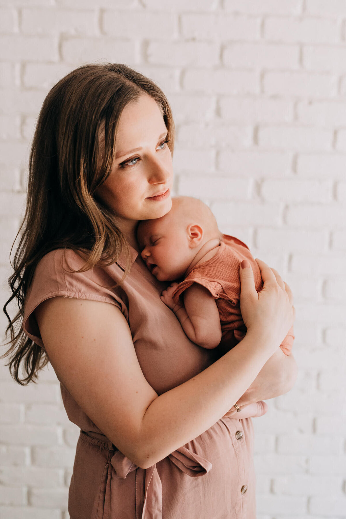 A young mother holds her newborn daughter standing in front of a white brick backdrop