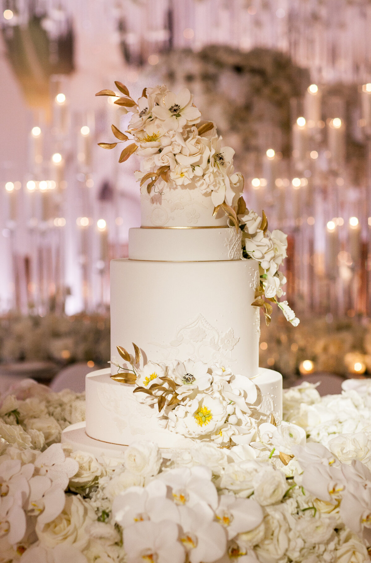 The photo showcases a breathtaking three-tiered white wedding cake adorned with intricate floral designs and clusters of white and gold flowers, elegantly captured at The New York Public Library in New York City by renowned photographer Christian Oth. Videography for this enchanting scene was expertly handled by East West Films. The exquisite cake is artfully complemented by a lavish arrangement of white roses and orchids, creating a stunning display against a backdrop of twinkling lights that adds a touch of enchantment to the scene.