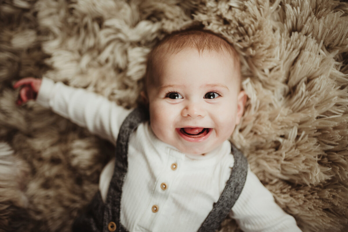 A baby boy gleefully smiles at the camera while lying on a fuzzy rug.