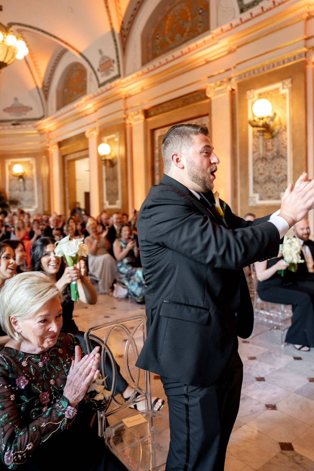 A man in a black suit stands clapping in an ornately decorated hall with marble floors. Seated guests also clap and smile. The atmosphere appears celebratory and joyful.