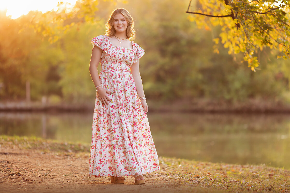Dallas senior photographer capturing a young woman in a floral dress, standing by a peaceful river during sunset.